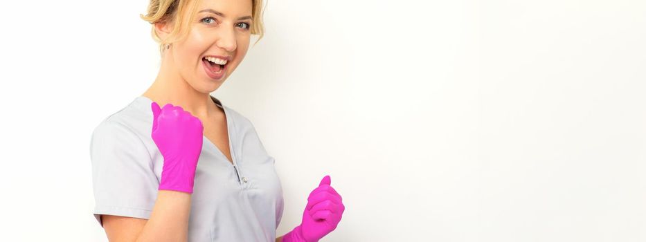 Happy caucasian woman doctor wearing pink gloves celebrates and raising fists on white background