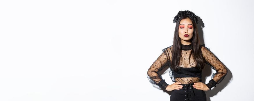 Image of beautiful asian girl in halloween costume close eyes and pouting, waiting for kiss, standing over white background wearing gothic black dress with wreath made of roses.