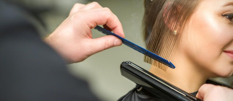 Hairstylist is straightening short hair of young brunette woman with a flat iron in a hairdresser salon, close up