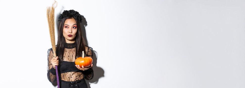 Image of sassy witch in gothic lace dress, holding broom and pumpkin, looking at upper left corner with halloween banner, white background.