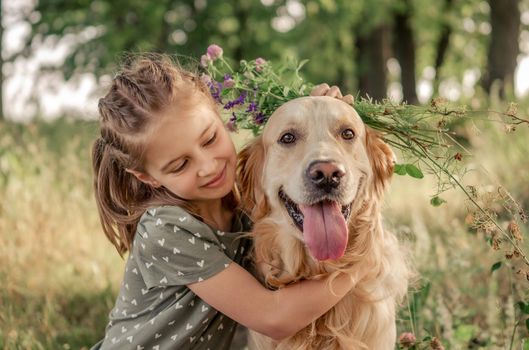 Portrait of beautiful preteen girl petting and hugging golden retriever dog looking at the camera outdoors. Kid with doggy pet in the field in summer time