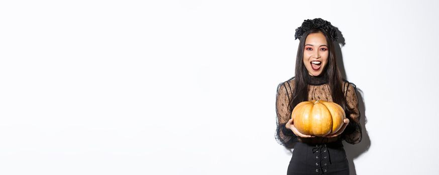 Image of joyful asian woman in witch costume, celebrating halloween, giving you pumpkin and looking scary, standing over white background.