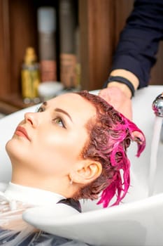 Washing dyed female hair. A young caucasian woman having her hair washed in a beauty salon. Professional hairdresser washes pink color paint off of a customer's hair
