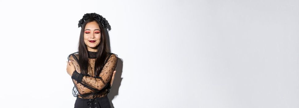 Happy cute asian woman hugging herself and smiling dreamy, wearing halloween costume of witch or widow, standing over white background.