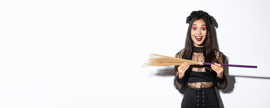 Excited young asian girl in witch costume looking amazed, showing small broom, standing over white background.