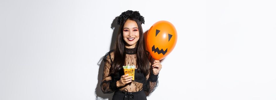 Image of cheerful asian girl trick or treating on halloween, holding sweets and orange balloon with scary face.