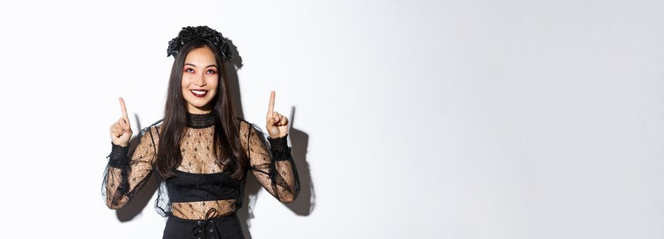 Beautiful asian woman in undead widow costume, wearing black lace dress for halloween, pointing fingers up and smiling pleased, looking at your logo, standing over white background.