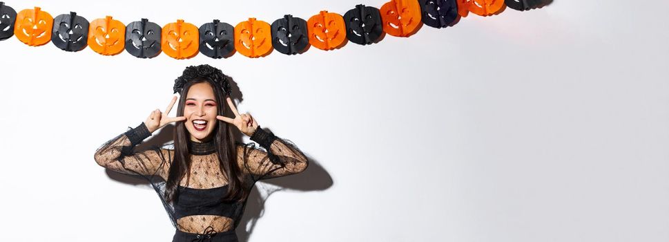 Cute happy asian woman enjoying halloween party, showing peace gesture and smiling, wearing witch costume, standing against pumpkin banners decoration.