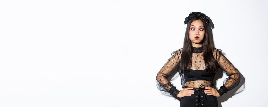 Portrait of funny and carefree asian girl having fun on halloween, making faces and holding breath, standing in gothic witch costume over white background.