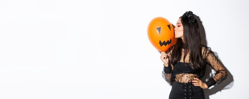 Stylish asian woman in gothic lace dress celebrating halloween, kissing orange balloon with face, standing over white background.