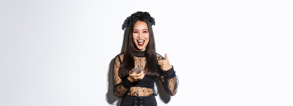 Portrait of satisfied asian woman in elegant gothic dress and black wreath showing thumbs-up while using mobile phone, standing over white background.