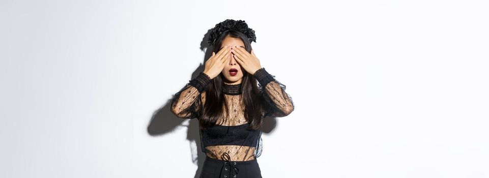 Hopeful and excited stylish asian girl in witch dress close her eyes and waiting for surprise, open mouth amused, wearing halloween costume, standing over white background.