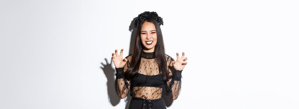 Sassy beautiful asian woman in black lace dress and wreath trying to scare you, raising hands up, acting like evil witch on halloween trick or treat event, standing over white background.