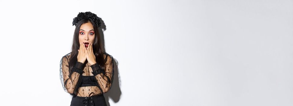 Surprised beautiful asian woman in witch costume looking amazed. Female wearing halloween gothic dress and wreath, open mouth aamused, standing over white background.