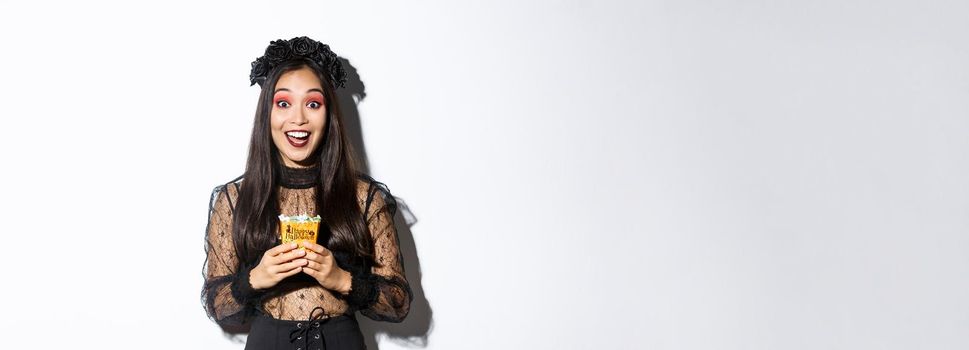 Beautiful asian girl smiling happy, holding sweets, wearing witch costume on halloween, enjoying trick or treating.