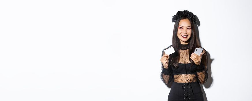 Image of beautiful asian woman in gothic lace dress and black wreath, looking aside pleased and smiling, holding mobile phone with credit card.