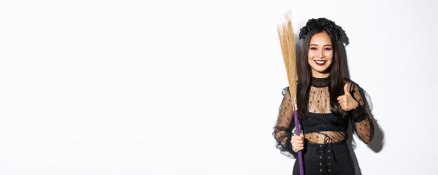 Image of smiling asian woman in witch costume with broom, showing thumbs-up in approval, standing over white background.