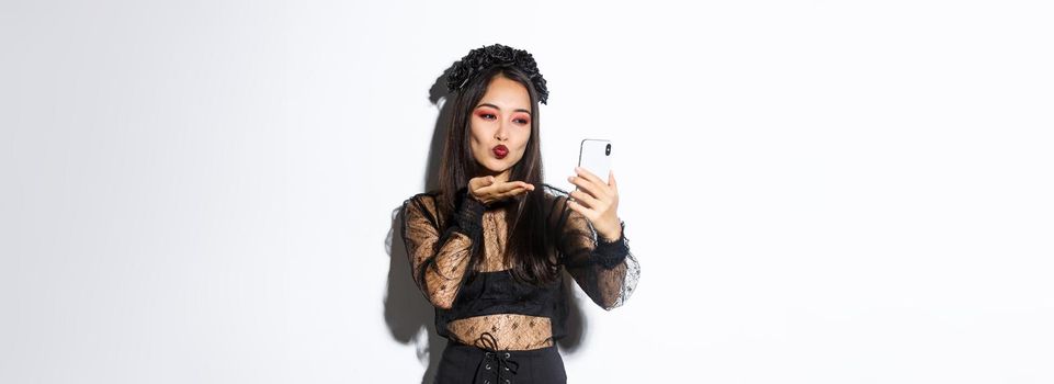 Portrait of stylish asian female blogger with gothic makeup and halloween costume sending air kiss at mobile phone camera, record video or having videocall, standing over white background.