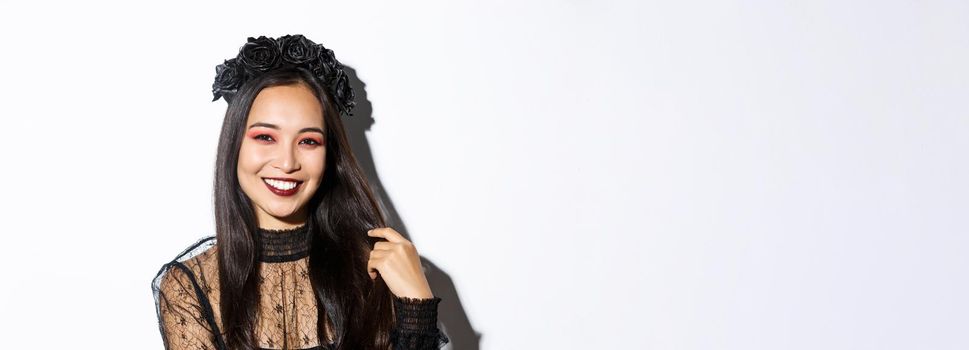 Close-up of beautiful elegant asian woman in black wreath and gothic lace dress smiling, standing over white background, dressed-up for halloween party.