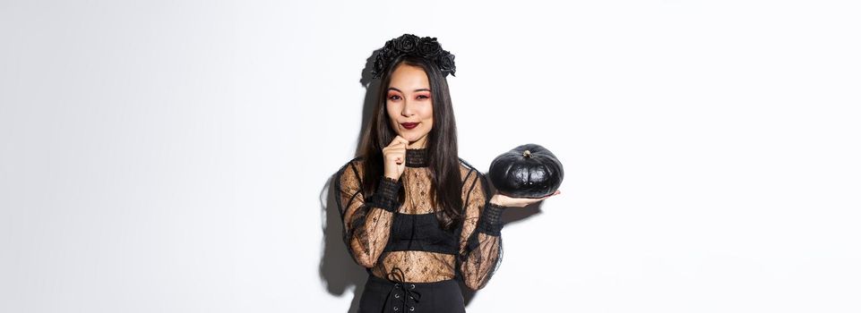 Image of thoughtful smiling asian witch, girl in halloween costume looking at camera and showing black pumpkin.