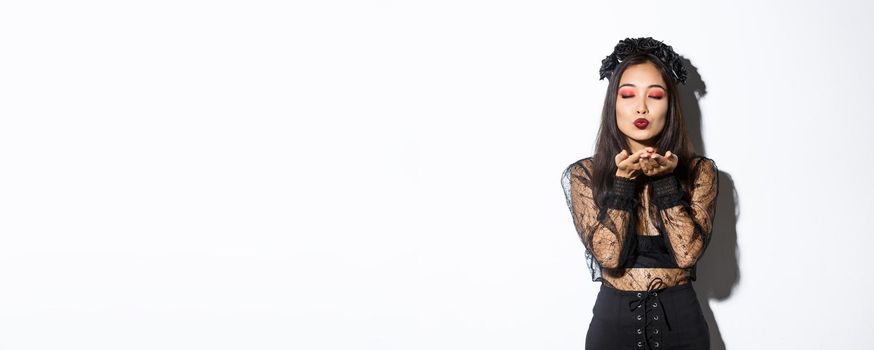 Image of beautiful and elegant asian woman in witch costume, close eyes and sending air kiss t camera, wearing gothic dress with black wreath, standing over white background.