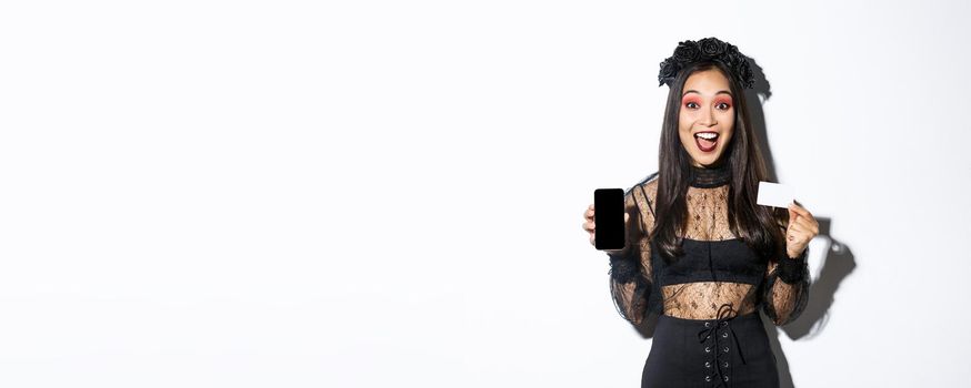 Cheerful beautiful asian woman in halloween costume, dressed as witch and showing smartphone with credit card, standing over white background.