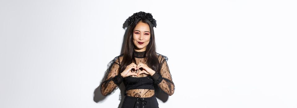 Image of lovely smiling asian woman love celebrating halloween, showing heart gesture and looking at camera, standing over white background in gothic lace dress with wreath.