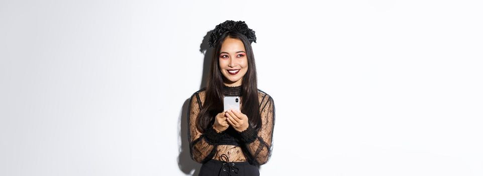 Portrait of cunning beautiful asian woman, witch in gothic lace dress using mobile phone, smiling and looking at upper left corner, standing over white background.