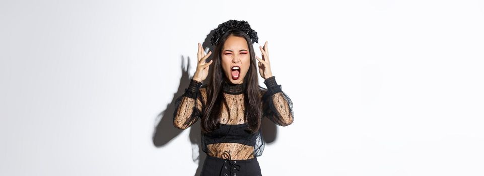 Image of angry asian woman in witch costume yelling at someone, looking mad, losing temper and screaming, standing over white background pissed-off.