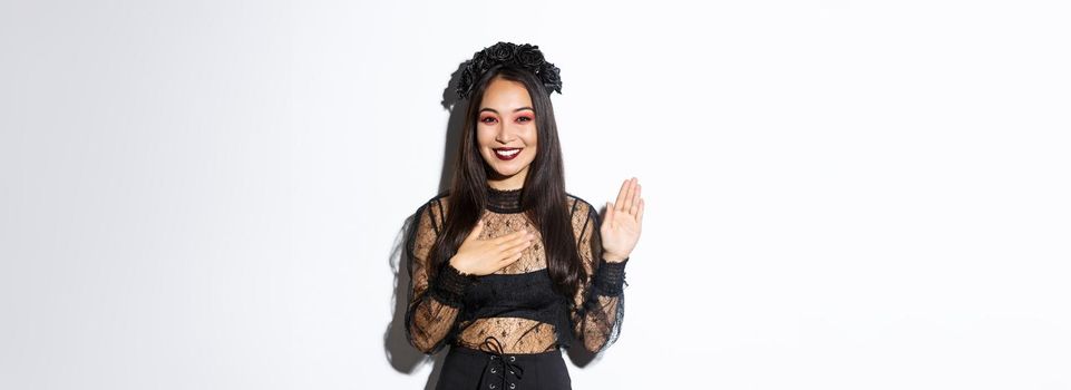 Image of cute sincere asian girl in halloween costume making promise, holding one hand on heart while swearing, being honest, standing over white background.