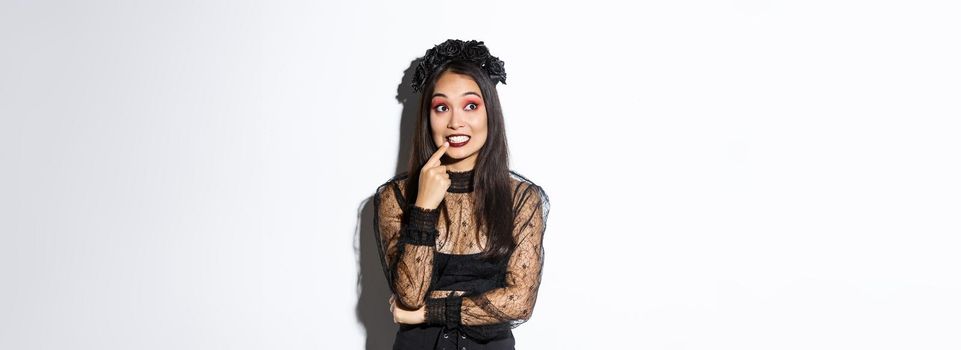 Image of intrigued young cute woman in witch dress, celebrating halloween, thinking about something interesting, looking upper left corner, standing over white background.