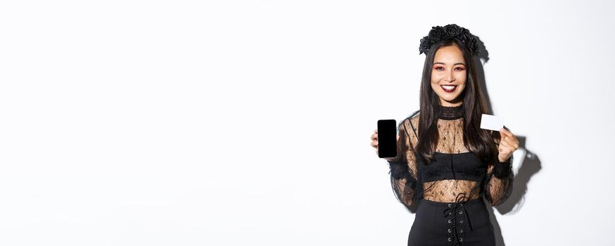 Smiling attractive woman in black gothic dress showing credit card and smartphone display, standing over white background in halloween costume.