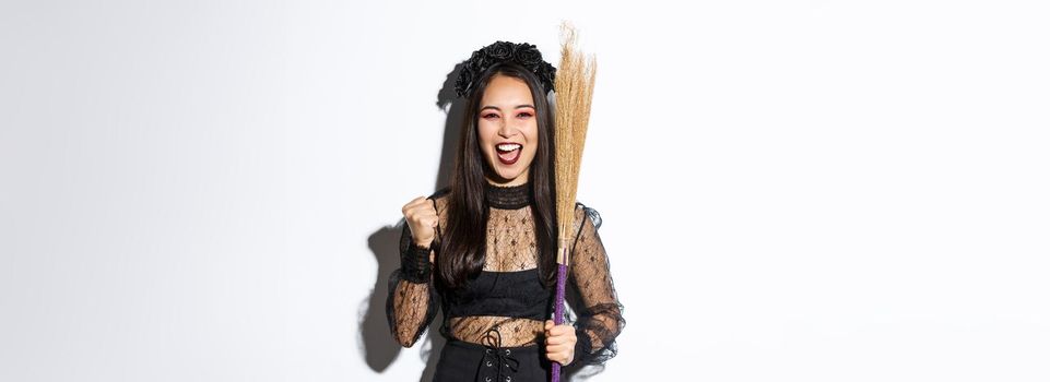 Image of cheerful asian girl in witch costume celebrating victory, holding broom, saying yes and raising fist in triumph, white background.