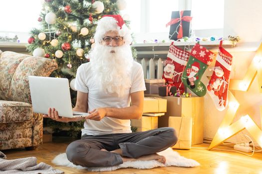Happy Santa Claus in suit sitting on an armchair with laptop on christmas eve. Online e-commerce store christmas vacation online sale concept. Against background of christmas tree and garlands on wall.