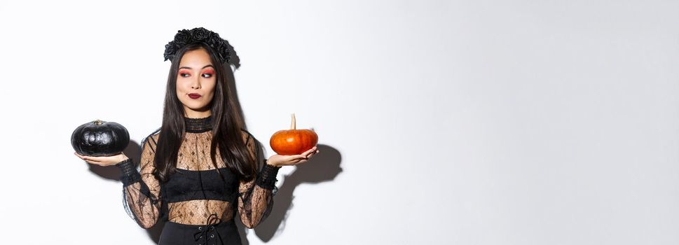 Thoughtful stylish woman in witch costume, picking pumpkin for halloween, making decision while standing over white background.