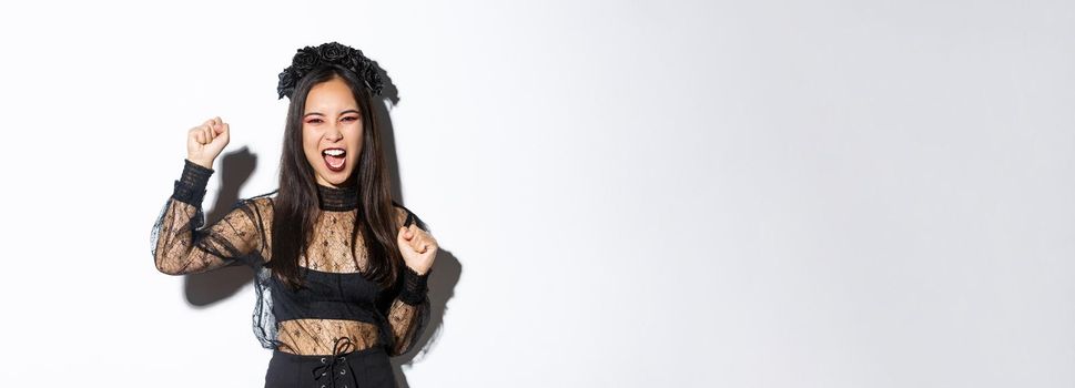 Excited asian woman in elegant gothic lace dress and wreath celebrating halloween, enjoying party, raising hand up and yelling joyful, standing over white background.