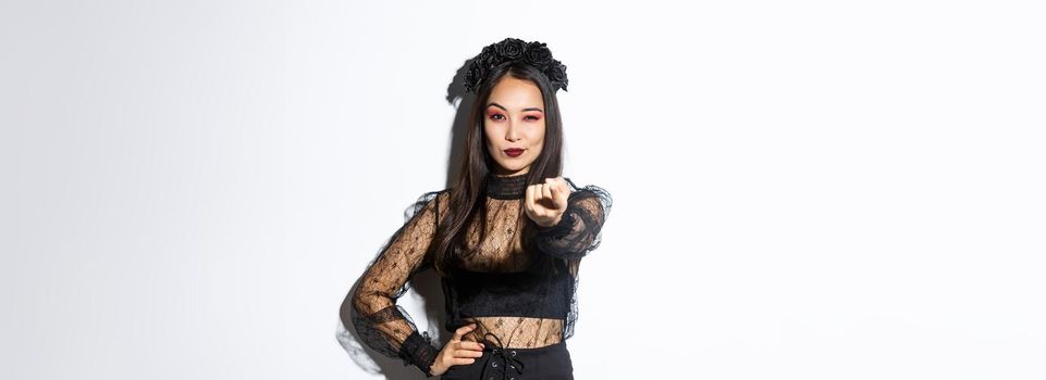Confident beautiful asian woman in sexy witch costume lure you, tell come closer with extended finger, celebrating halloween and wearing gothic dress, standing over white background.