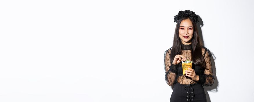 Smiling cute asian woman celebrating halloween, holding sweets and grinning happy, trick or treating in witch costume.