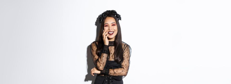 Image of happy good-looking asian woman in gothic dress and black wreath, laughing at halloween party, standing over white background.