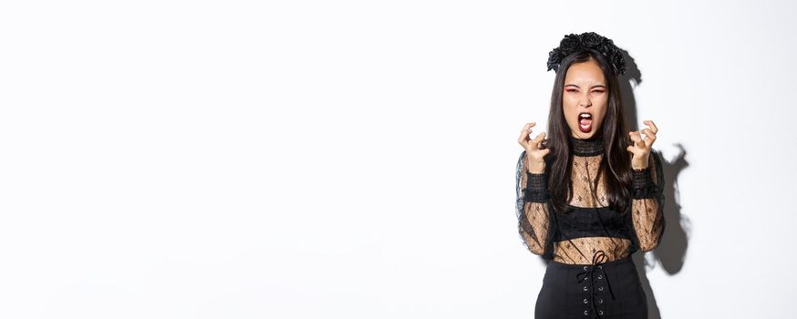 Image of mad woman in gothic lace dress and black wreath yelling angry and clenching fists, standing in halloween costume with furious expression, standing over white background.