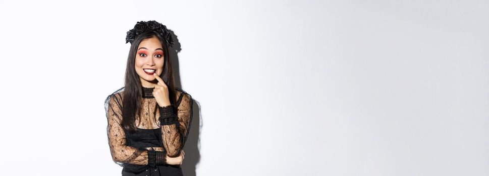 Image of thoughtful beautiful asian woman in halloween party costume looking amused and intrigued, standing over white background wearing gothic dress and wreath.