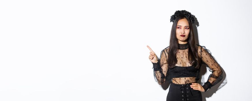 Unamused and skeptical asian beautiful woman in witch dress, looking at upper left corner with displeased smirk, standing over white background, showing logo or promo banner.