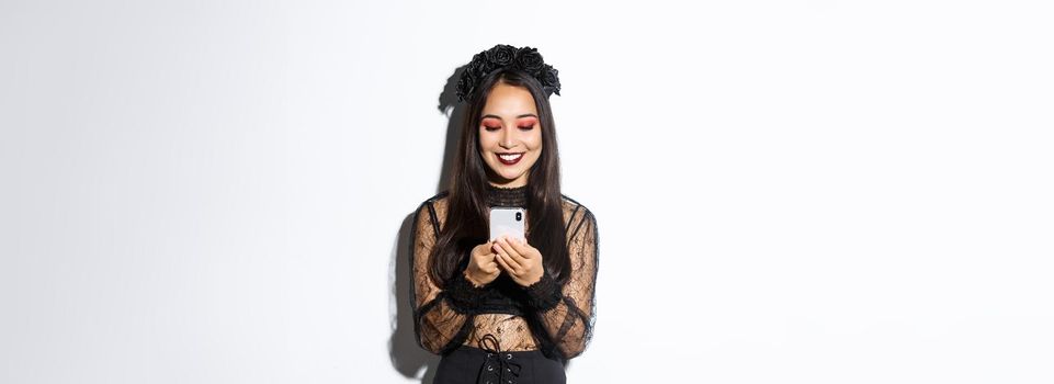 Image of beautiful asian woman in gothic lace dress impersonating witch on halloween party, standing with smartphone and smiling over white background.