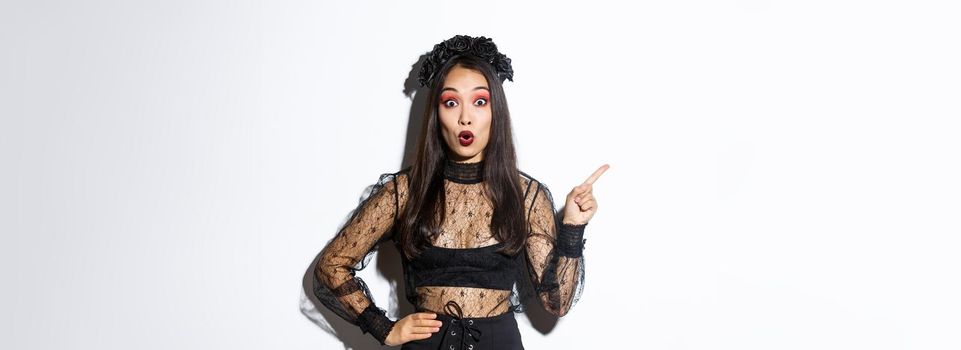 Surprised attractive asian woman in witch outfit pointing finger at upper left corner, showing halloween advertisement, promo of party. Beautiful female in black gothic dress looking amazed.