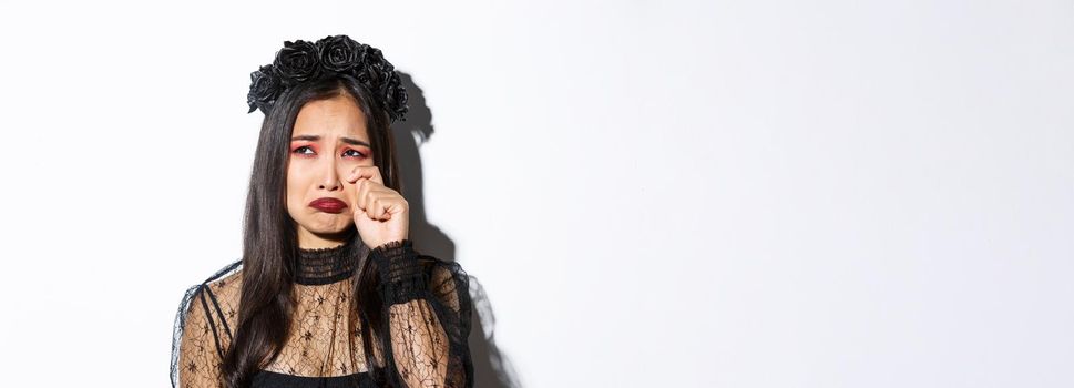 Close-up of young asian woman in witch costume crying and looking miserable, feeling sad, standing over white background.