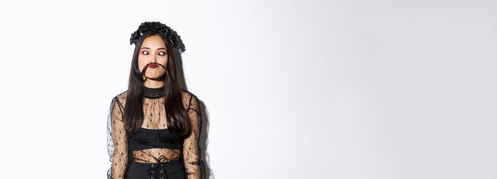 Portrait of silly and funny asian girl foolind around, grimacing and making moustache from hair strand, standing over white background in halloween costume.