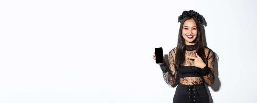 Happy beautiful asian girl in witch costume pointing finger at smartphone screen with pleased smile, showing halloween announcement, white background.