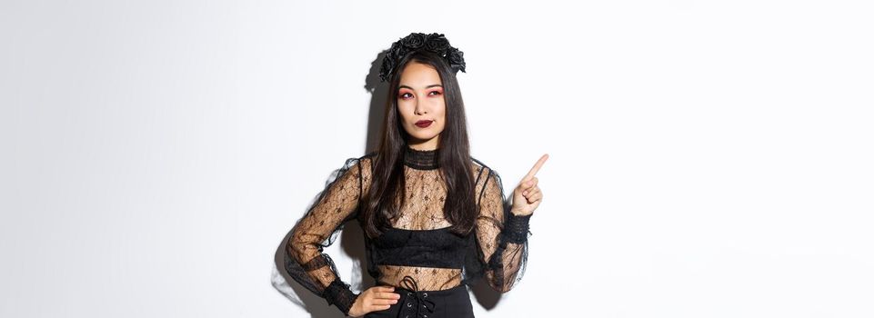 Curious attractive asian woman in witch costume, pointing finger upper left corner, looking interested in halloween promo, standing over white background and thinking.