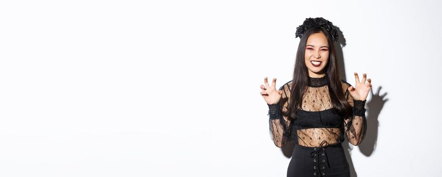 Sassy beautiful asian woman in black lace dress and wreath trying to scare you, raising hands up, acting like evil witch on halloween trick or treat event, standing over white background.