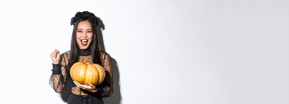 Image of excited beautiful asian woman celebrating halloween, wearing witch outfit and holding pumpkin, yelling with rejoice.